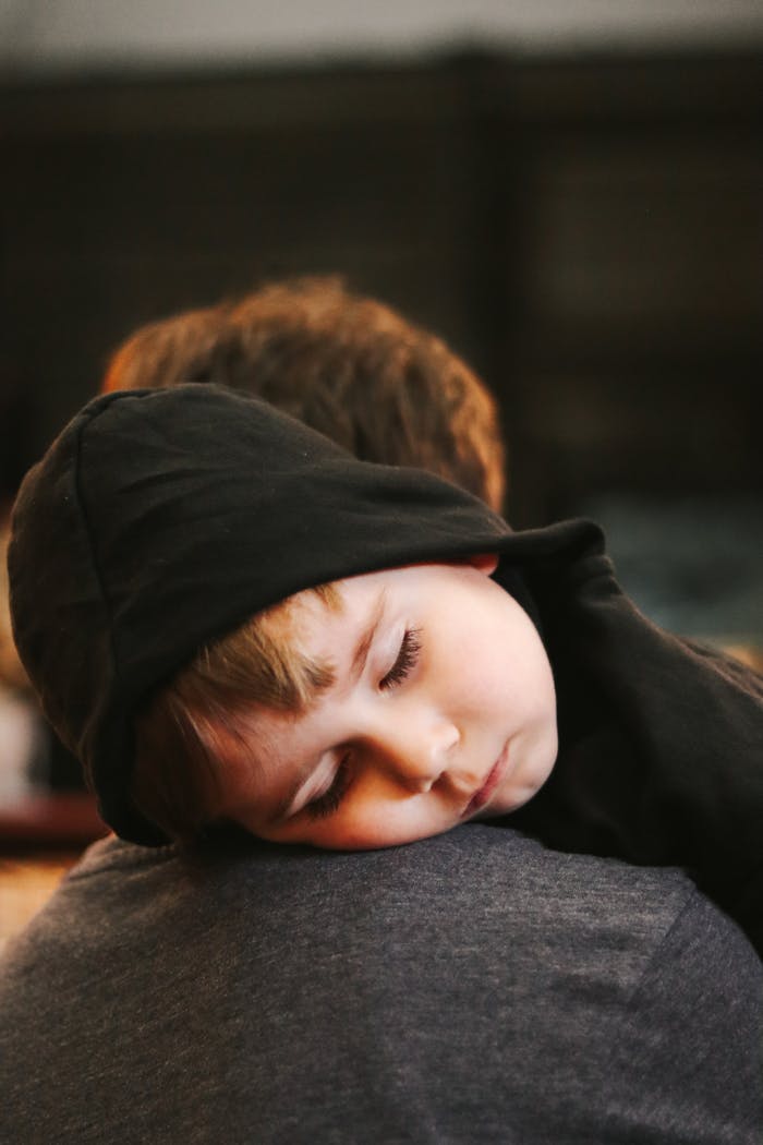 gallery-5 A young child in a hoodie peacefully sleeping on a parent's shoulder indoors.