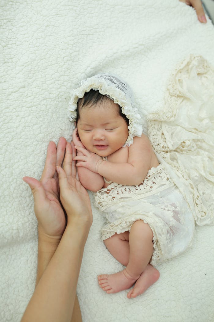 gallery-1 Charming newborn baby in lace bonnet and wrap sleeping peacefully, supported gently by adult hands.